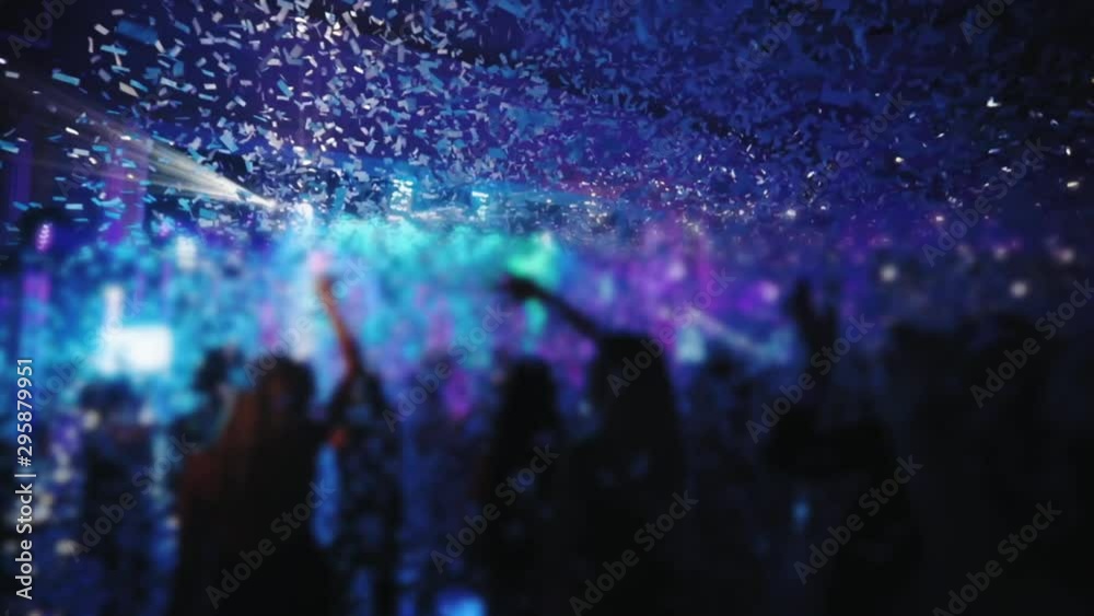 people jump with raised hands under white confetti fall against blurry illuminated stage in night club slow motion
