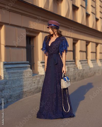 Full length outdoor portrait of young beautiful elegant woman in long blue evening dress and purple hat with gray leather handbag standing and posing at old city street on a sunny evening day