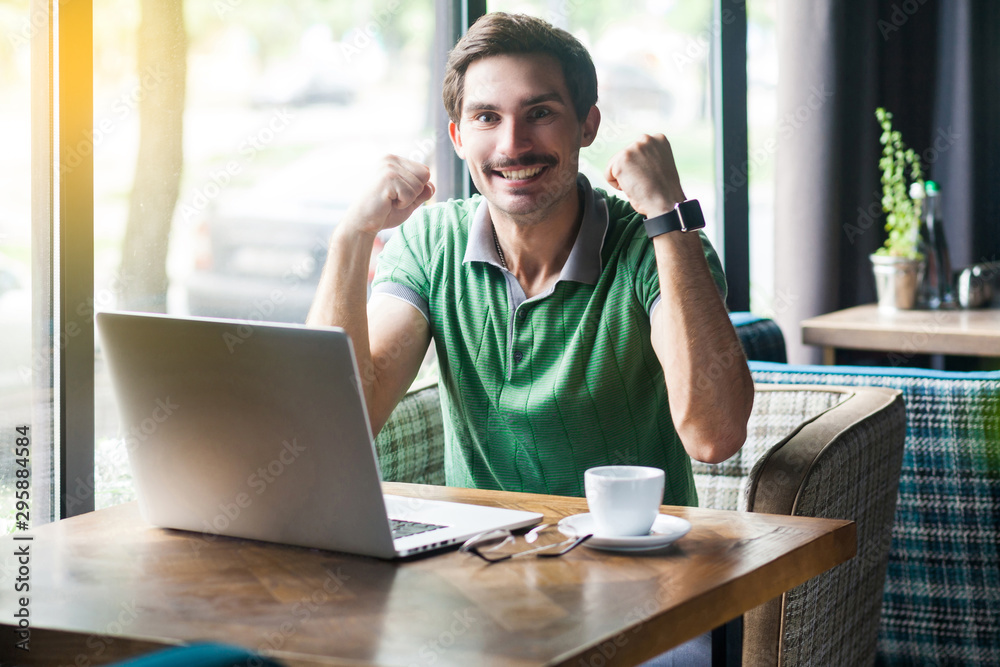 Fototapeta premium Yes i win! Young happy successful excited businessman in green t-shirt sitting with laptop and celebrating his victory and success. business and freelancing concept. indoor shot near window at daytime