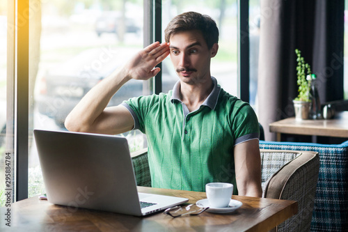 Фотография Yes sir! Young businessman in green t-shirt sitting and looking at laptop display on video call with salute gesture and ready to complete the mission