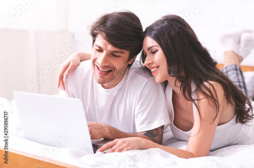 Young married couple lying on bed at home and using laptop