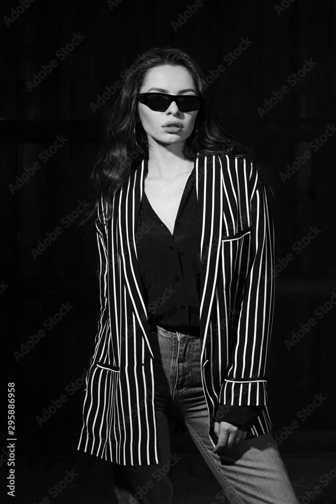 Naklejka premium Body length interior portrait of stylish young female modelwearing grey jeans and striped blazer standing and posing in dark room. Vogue style. Studio shot