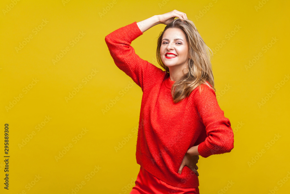 Portrait of sensual happy blonde woman with red lipstick in casual sweater standing, touching hair with hand and looking at camera, fashion model. indoor studio shot isolated on yellow background Portrait of sensual happy blonde woman with red lipstick in casual sweater standing, touching hair with hand and looking at camera, fashion model. indoor studio shot isolated on yellow background