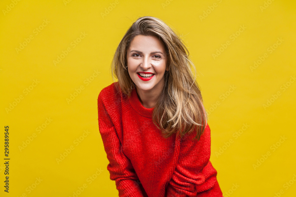 Portrait of positive blonde woman with red lipstick in bright casual sweater standing, looking at camera with joyful and charming smile, enjoying life. indoor studio shot isolated on yellow background Portrait of positive blonde woman with red lipstick in bright casual sweater standing, looking at camera with joyful and charming smile, enjoying life. indoor studio shot isolated on yellow background