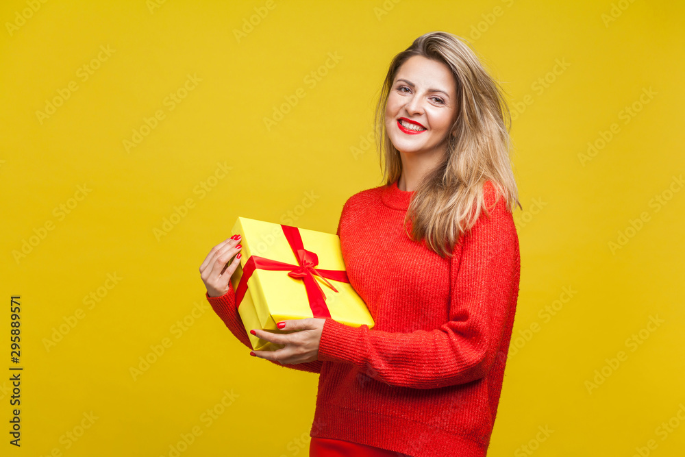 Portrait of delighted beautiful blonde woman with red lipstick in bright casual sweater, holding wrapped gift box and looking at camera, toothy smile. indoor studio shot isolated on yellow background Portrait of delighted beautiful blonde woman with red lipstick in bright casual sweater, holding wrapped gift box and looking at camera, toothy smile. indoor studio shot isolated on yellow background