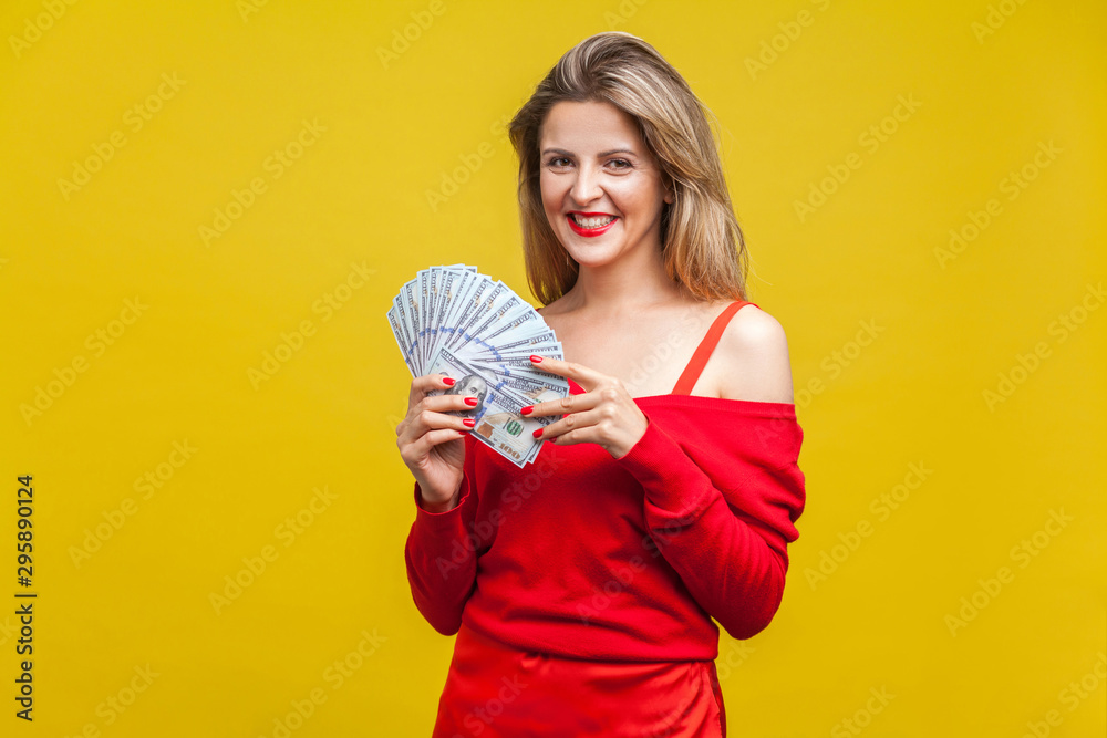 Portrait of wealthy beautiful young woman in red dress standing with dollar bills, looking at camera and smiling excitedly, enjoying rich life. indoor studio shot isolated on yellow background Portrait of wealthy beautiful young woman in red dress standing with dollar bills, looking at camera and smiling excitedly, enjoying rich life. indoor studio shot isolated on yellow background