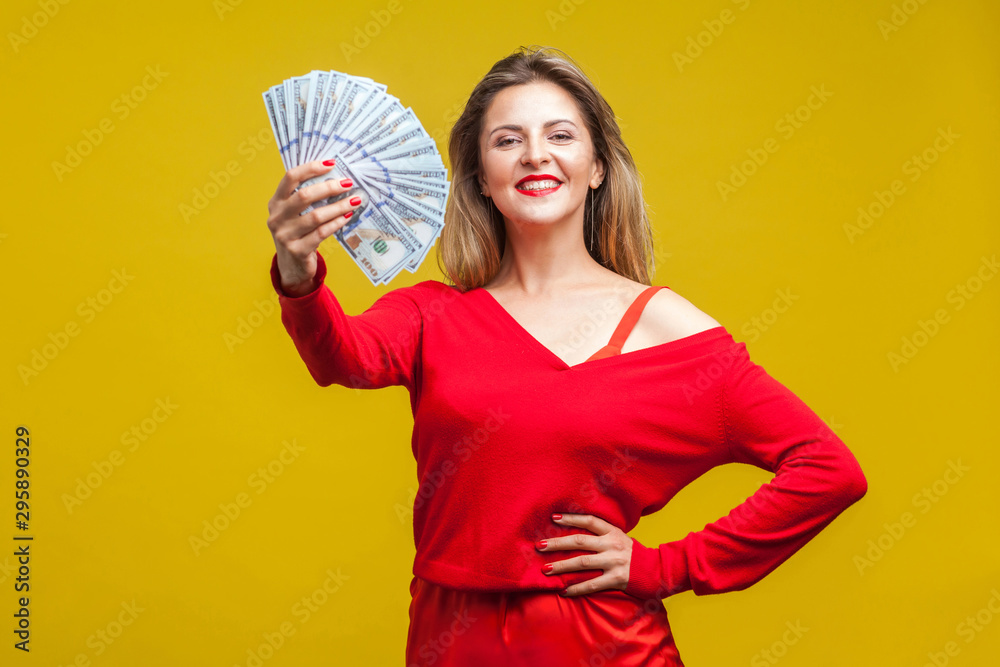 Portrait of arrogant wealthy beautiful woman in red dress standing showing dollar bills, looking at camera with smile, proud of big money, income. indoor studio shot isolated on yellow background Portrait of arrogant wealthy beautiful woman in red dress standing showing dollar bills, looking at camera with smile, proud of big money, income. indoor studio shot isolated on yellow background