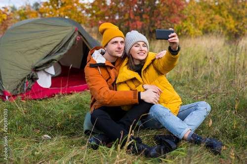 travel, love and autumn concept - happy couple in love sitting near green tent and taking selfie photo in autumn forest