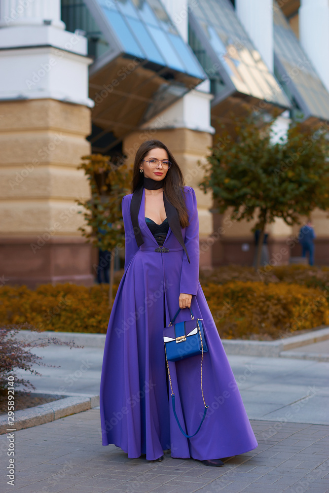 Young beautiful caucasian woman with long straight hair and makeup in purple trousers and long coat cape standing and posing at city street in autumn Young beautiful caucasian woman with long straight hair and makeup in purple trousers and long coat cape standing and posing at city street in autumn