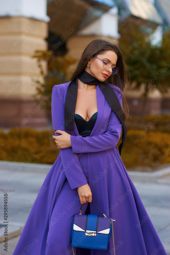 Young beautiful caucasian woman with long straight hair and makeup in purple trousers and long coat cape standing and posing at city street in autumn Young beautiful caucasian woman with long straight hair and makeup in purple trousers and long coat cape standing and posing at city street in autumn