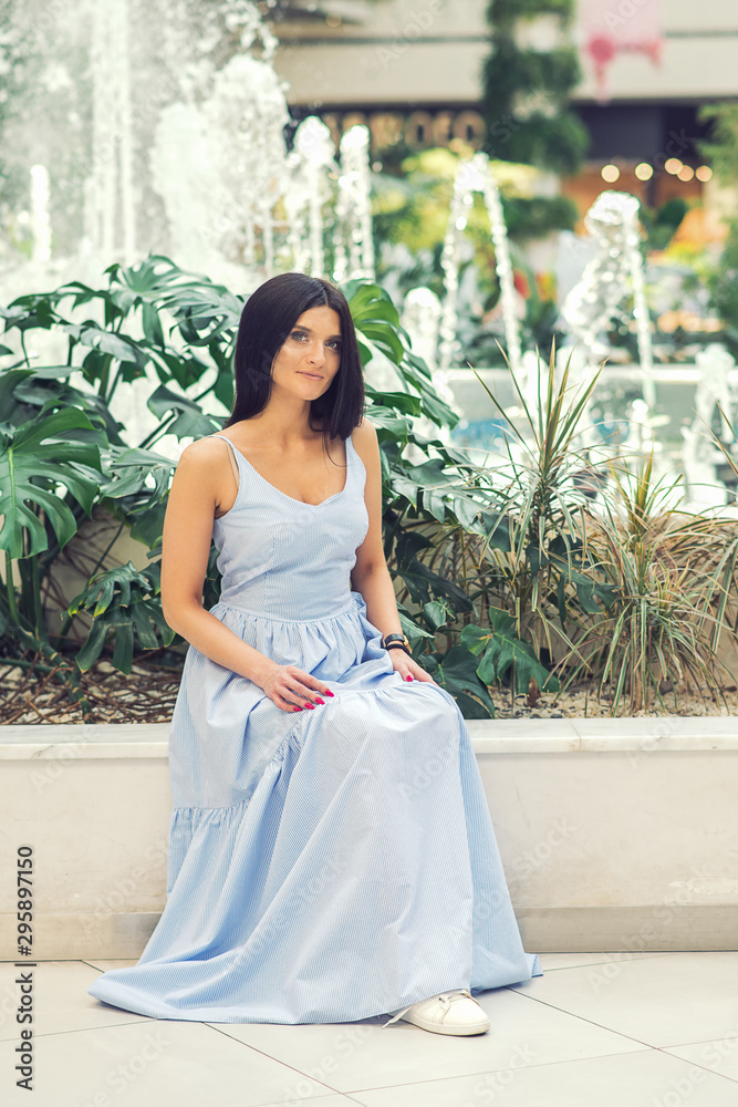 A young brunette woman sits near a fountain. Outdoor romantic portrait of attractive brunette woman wearing blue long dress is posing by fountain at park.