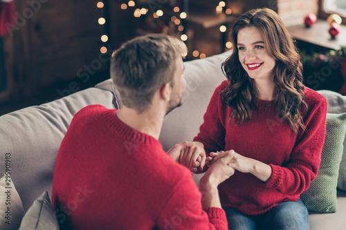 Behind view photo of emotional couple holding arms spending x-mas eve together in garland lights room sitting comfy sofa indoors wearing red sweaters