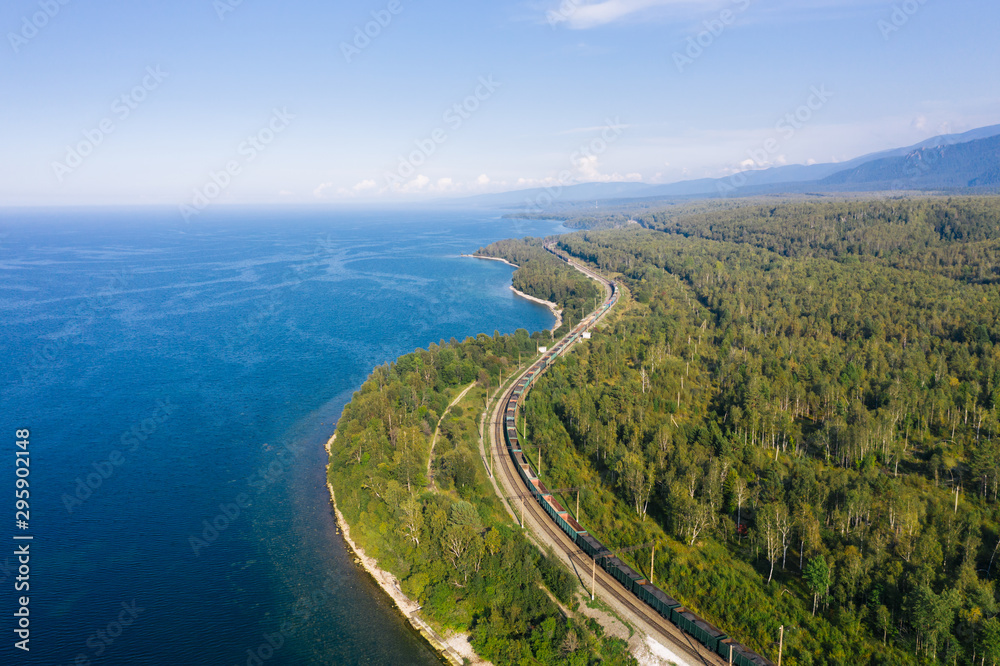 Aerial view of a freight train on the railroad of Trans-Siberian Railway on the shore of Baikal Lake with green forest trees in a sunny summer day. East Siberian Railway in Buryatia, Siberia, Russia