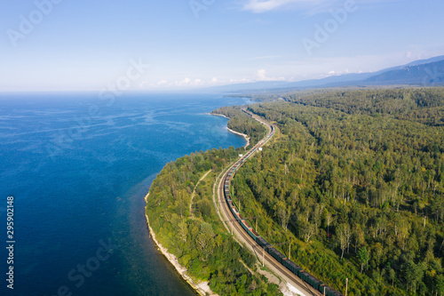 Aerial view of a freight train on the railroad of Trans-Siberian Railway on the shore of Baikal Lake with green forest trees in a sunny summer day. East Siberian Railway in Buryatia, Siberia, Russia © Vitaliy Kaplin