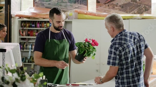 Busy young man working as florist in flower shop. Sales manager at cashier desk talking to client buying plant. Customer paying with credit card in store and speaking to business owner photo