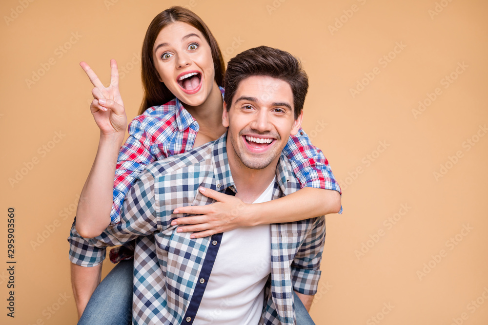 Photo of cheerful positive excited crazy boyfriend and girlfriend with her showing you v-sign with excited facial expression and him holding her piggyback isolated over beige pastel color background Photo of cheerful positive excited crazy boyfriend and girlfriend with her showing you v-sign with excited facial expression and him holding her piggyback isolated over beige pastel color background