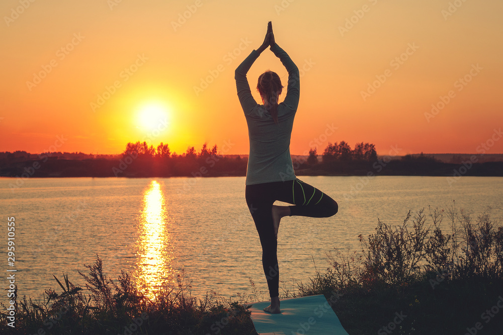 A young girl is standing on the lake at sunset, doing yoga. Stands in a pose of tree a Sathi Yoga. Balance, harmony, balance, concentration, relaxation.