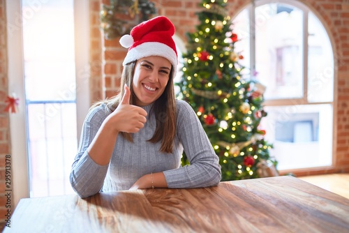 Young beautiful woman wearing santa claus hat at the table at home around christmas decoration doing happy thumbs up gesture with hand. Approving expression looking at the camera with showing success.