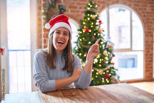 Young beautiful woman wearing santa claus hat at the table at home around christmas decoration with a big smile on face, pointing with hand and finger to the side looking at the camera.