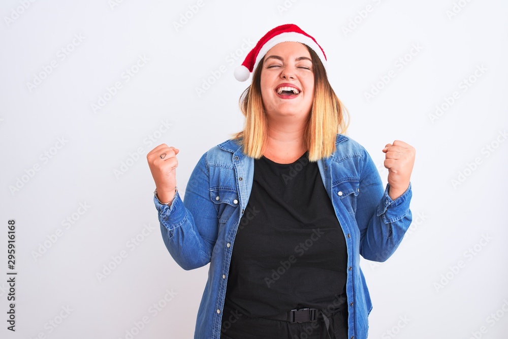 Young beautiful woman wearing Christmas Santa hat standing over isolated white background very happy and excited doing winner gesture with arms raised, smiling and screaming for success Young beautiful woman wearing Christmas Santa hat standing over isolated white background very happy and excited doing winner gesture with arms raised, smiling and screaming for success