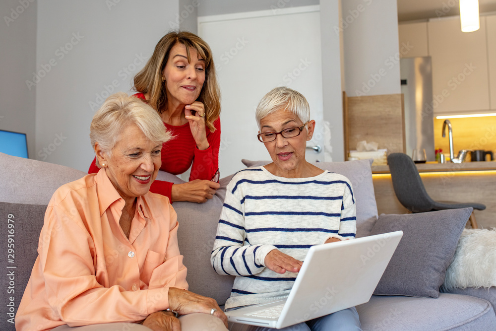 Browsing some old photos on the laptop. Smiling friends looking at laptop together and eating cookies at home on couch. Portrait of 3 senior female friends at home. Browsing some old photos on the laptop. Smiling friends looking at laptop together and eating cookies at home on couch. Portrait of 3 senior female friends at home.