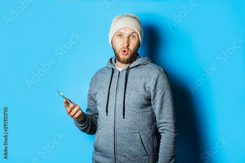 Man with a surprised face in a hoodie and hat is holding a phone on a blue background. Shock, surprise, mobile app