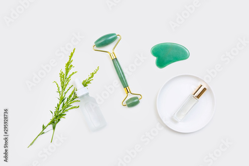 Gua sha stone, jade roller, clear perfume, white glass eye dropper serum bottle / still life arranged with green wildflowers and isolated on white surface / natural clean beauty concept