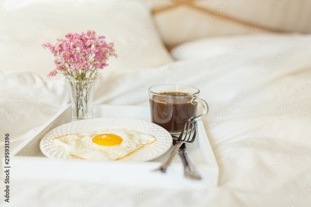 White tray with breakfast on a bed in a hotel room. Fried egg, cup of coffee and flowers in white sheets in light bedroom. White tray with breakfast on a bed in a hotel room. Fried egg, cup of coffee and flowers in white sheets in light bedroom.