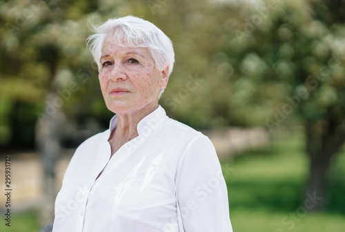 Elegant elderly gray-haired woman in the white shirt is standing in a park on a warm sunny day
