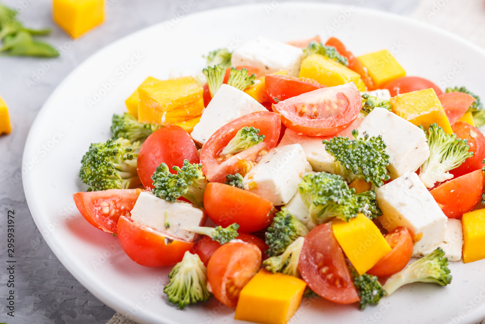 Vegetarian salad with broccoli, tomatoes, feta cheese, and pumpkin on white ceramic plate on a gray concrete background, side view, close up.
