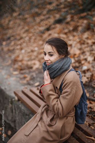 Autumn outdoor portrait of beautiful young woman in autumn park, sitting on a bench and wraps in warm knitted scarf.. © vitaliymateha