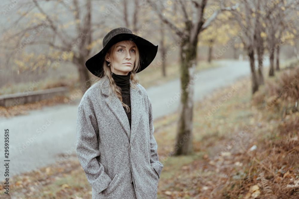 Young caucasian woman in a hat and a gray coat in the rain against the backdrop of an autumn landscape. Pensive melancholy autumn mood.