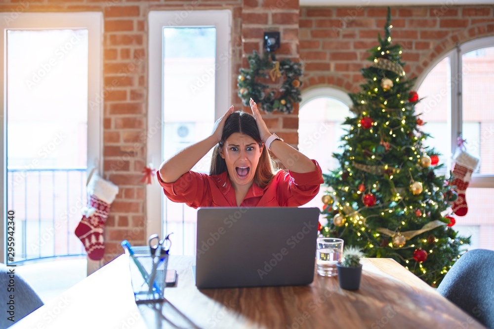 Beautiful woman sitting at the table working with laptop at home around christmas tree Crazy and scared with hands on head, afraid and surprised of shock with open mouth Beautiful woman sitting at the table working with laptop at home around christmas tree Crazy and scared with hands on head, afraid and surprised of shock with open mouth