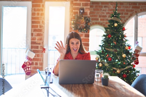 Beautiful woman sitting at the table working with laptop at home around christmas tree Waiving saying hello happy and smiling, friendly welcome gesture