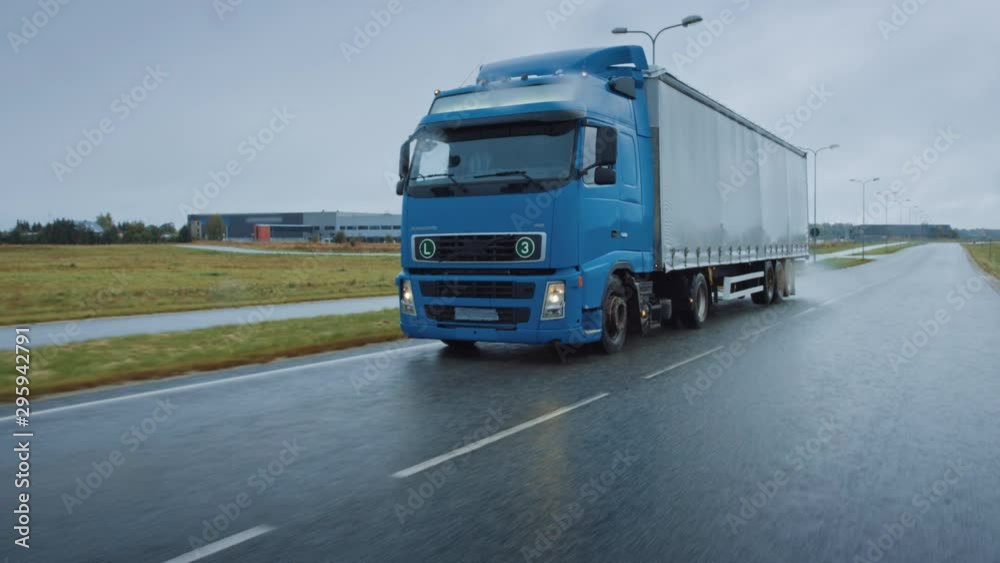 Long Haul Semi-Truck with Cargo Trailer Full of Goods Travels on the Highway Road. Daytime Driving Across Continent Through Rain. Industrial Retail Warehouses Area. Front View Following Shot Long Haul Semi-Truck with Cargo Trailer Full of Goods Travels on the Highway Road. Daytime Driving Across Continent Through Rain. Industrial Retail Warehouses Area. Front View Following Shot