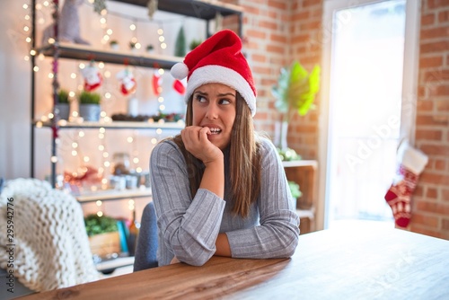 Young beautiful woman wearing christmas hat sitting at the table at home looking stressed and nervous with hands on mouth biting nails. Anxiety problem.
