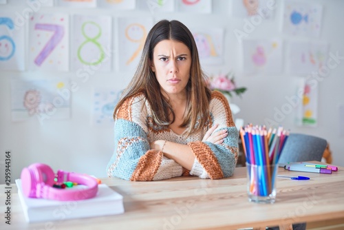 Young beautiful teacher woman wearing sweater and glasses sitting on desk at kindergarten skeptic and nervous, disapproving expression on face with crossed arms. Negative person.