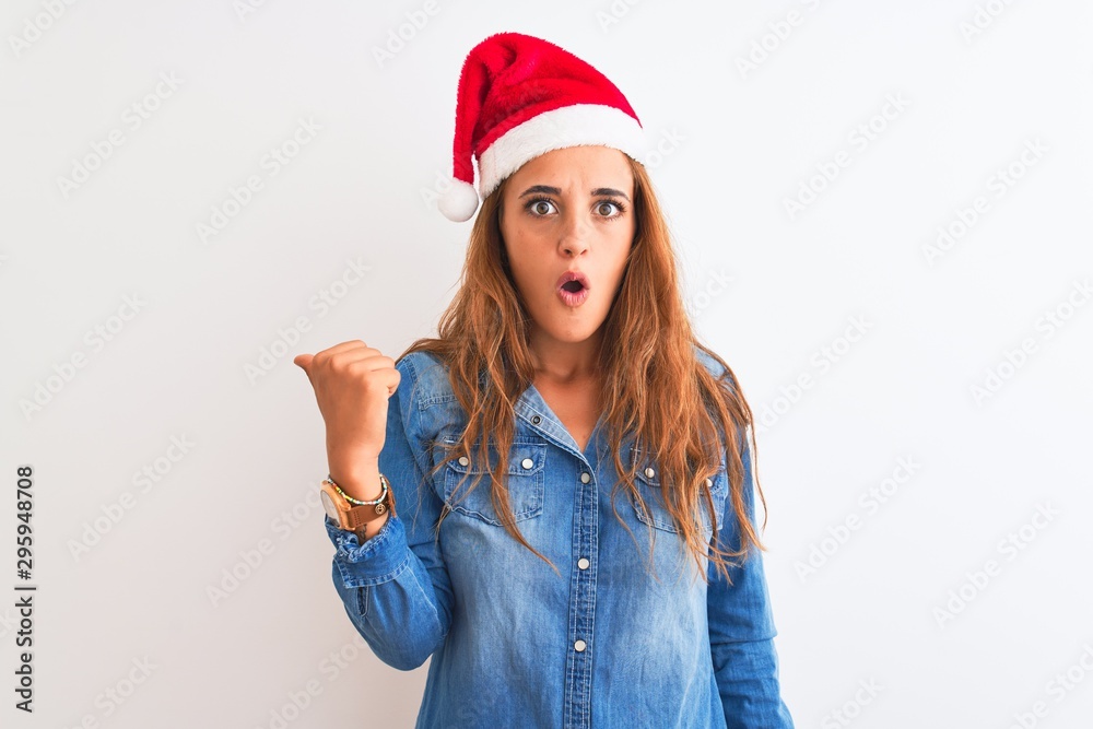 Young beautiful redhead woman wearing christmas hat over isolated background Surprised pointing with hand finger to the side, open mouth amazed expression. Young beautiful redhead woman wearing christmas hat over isolated background Surprised pointing with hand finger to the side, open mouth amazed expression.