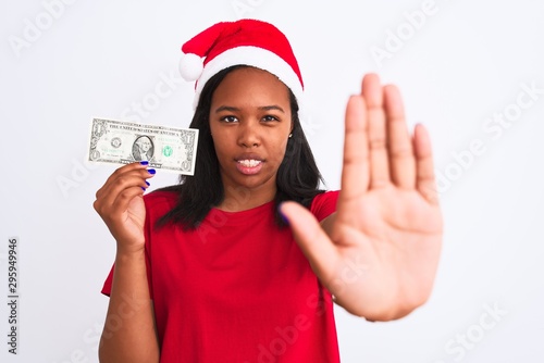 Young african american woman wearing christmas hat and holding one dollar banknote with open hand doing stop sign with serious and confident expression, defense gesture © Krakenimages.com