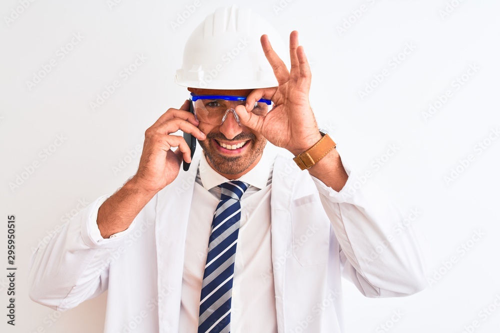 Young scientist man wearing safety helmet and calling using smartphone over isolated background with happy face smiling doing ok sign with hand on eye looking through fingers Young scientist man wearing safety helmet and calling using smartphone over isolated background with happy face smiling doing ok sign with hand on eye looking through fingers