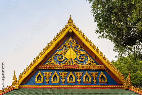 Bang Saen, Thailand - March 16, 2019: Wang Saensuk Buddhist Monastery. Gold, maroon and blue facade decoration op main open prayer hall against silver sky and under green foliage. photo