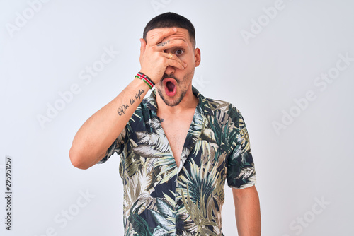 Young handsome man wearing Hawaiian summer shirt over isolated background peeking in shock covering face and eyes with hand, looking through fingers with embarrassed expression.