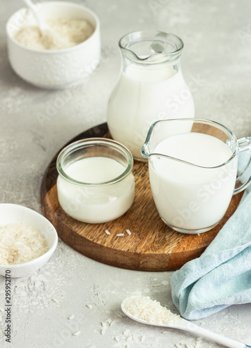 Rice milk in jugs and rice in a white ceramic bowls and spoon on a light grey stone background. Alternative type of milk. Vegan substitute dairy milk.