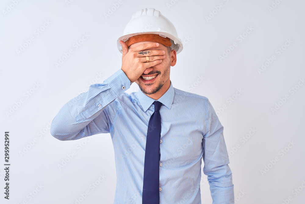 Young business man wearing contractor safety helmet over isolated background smiling and laughing with hand on face covering eyes for surprise. Blind concept. Young business man wearing contractor safety helmet over isolated background smiling and laughing with hand on face covering eyes for surprise. Blind concept.
