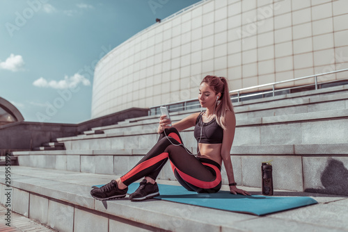 Athlete girl resting in summer in city on yoga mat. Rest after workout with fitness outdoor workout. In hands of phone  online application Internet  listens to music headphones. Free space for text.