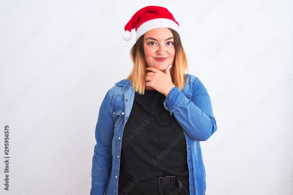 Young beautiful woman wearing Christmas Santa hat standing over isolated white background looking confident at the camera with smile with crossed arms and hand raised on chin. Thinking positive. Young beautiful woman wearing Christmas Santa hat standing over isolated white background looking confident at the camera with smile with crossed arms and hand raised on chin. Thinking positive.