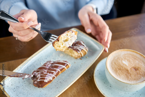 A young woman drinks coffee cappuccino in a restaurant and eats dessert. Traditional french eclairs with chocolate. Cake decorated with vanilla cream and chocolate frosting.