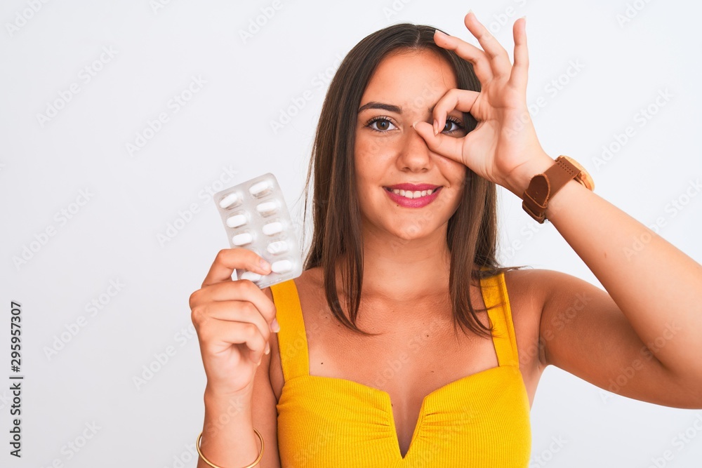 Young beautiful girl holding medicine pills standing over isolated white background with happy face smiling doing ok sign with hand on eye looking through fingers Young beautiful girl holding medicine pills standing over isolated white background with happy face smiling doing ok sign with hand on eye looking through fingers