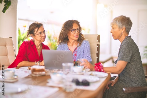 Meeting of middle age women having lunch and drinking coffee. Mature friends smiling happy using laptop at home on a sunny day