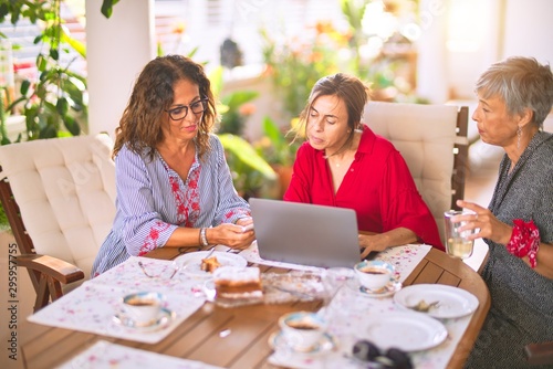 Meeting of middle age women having lunch and drinking coffee. Mature friends smiling happy using laptop at home on a sunny day